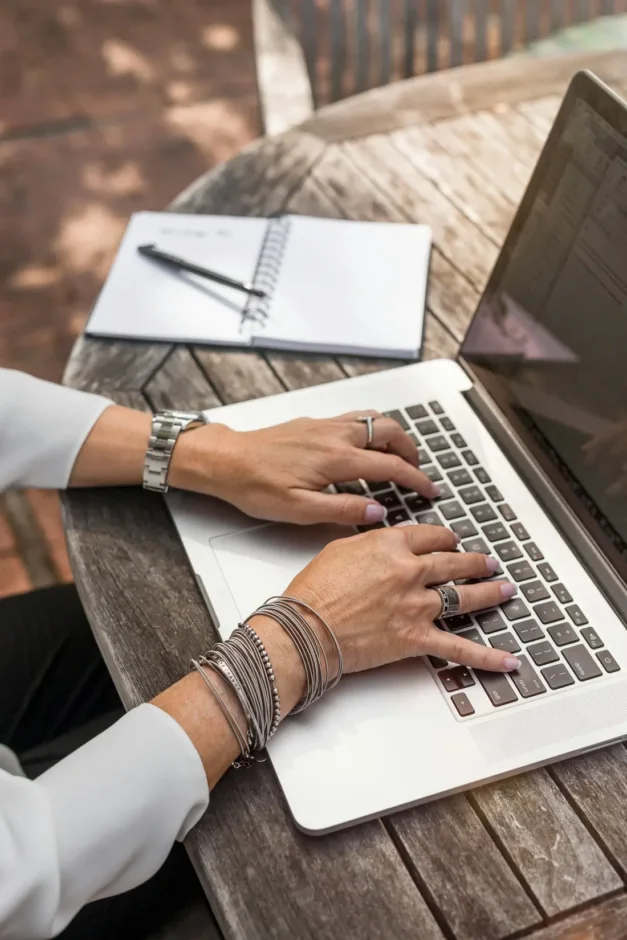 Mâini care scriu la un laptop, lângă un caiet